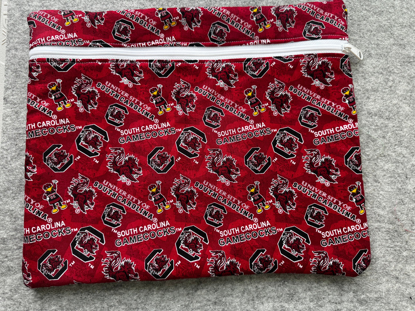 A folded fabric item, likely a pouch or sleeve, featuring a repeating South Carolina Gamecocks print in red, black, white, and yellow on a gray textured background.