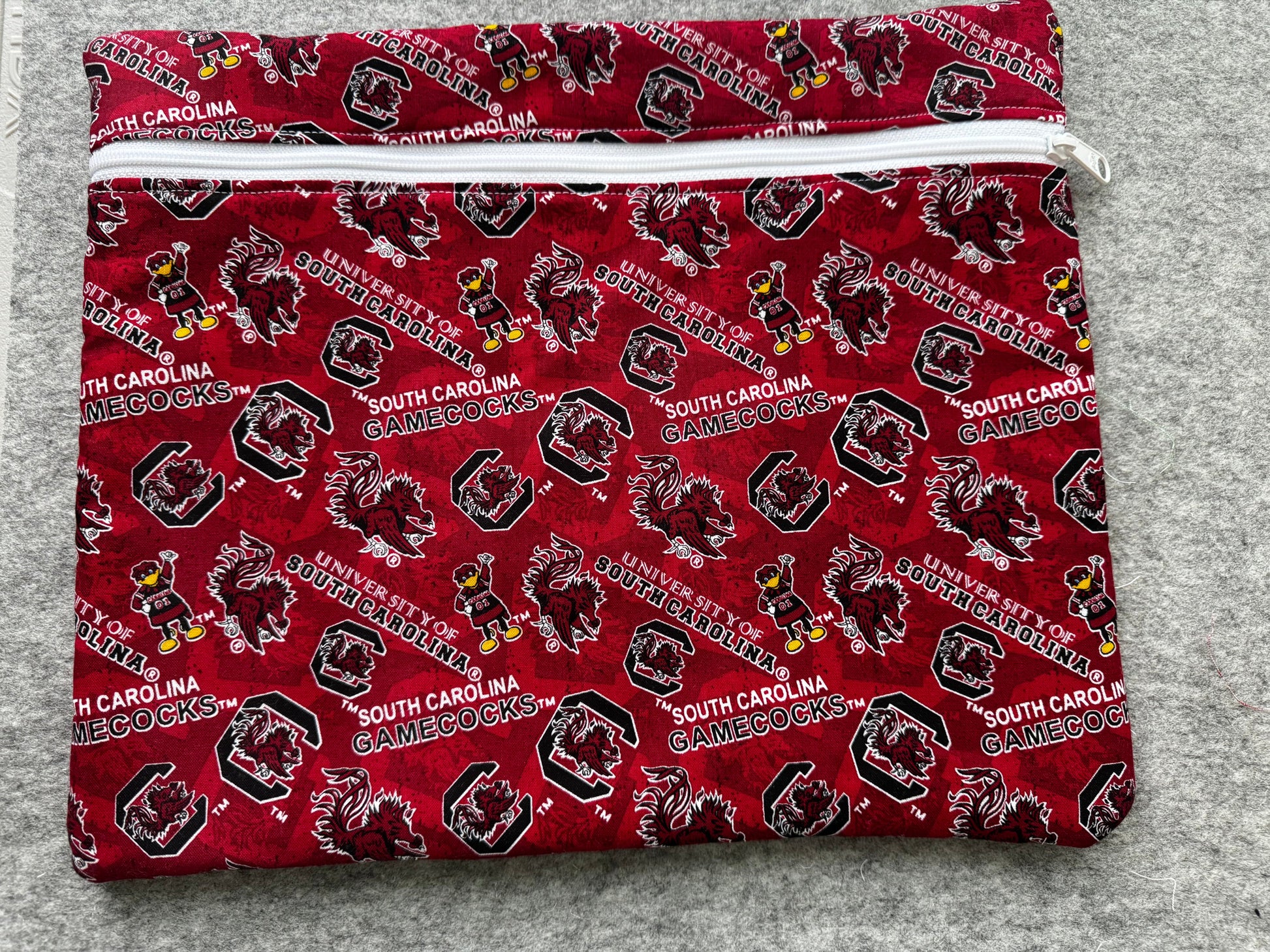 A folded fabric item, likely a pouch or sleeve, featuring a repeating South Carolina Gamecocks print in red, black, white, and yellow on a gray textured background.