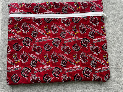 A folded fabric item, likely a pouch or sleeve, featuring a repeating South Carolina Gamecocks print in red, black, white, and yellow on a gray textured background.