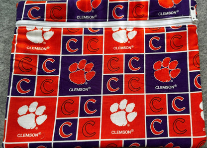 A folded fabric item, likely a pouch or small bag, featuring a repeating Clemson Tigers block pattern in red, purple, and white, laid flat on a gray textured background.
